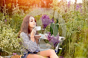 Young girl holds white cup with summer flowers and herbs on a picnic in the summer garden. Healing herbs and flowers. Health and