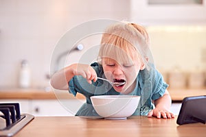 Young Girl At Home At Kitchen Counter Eating Breakfast Cereal From Bowl Whilst Watching Mobile Phone