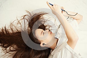 A young girl at home is lying on a bed with her hair spread out, looking at her smartphone, listening to music, taking selfies for