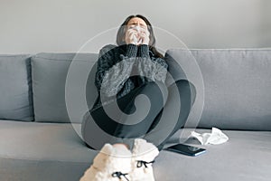 Young girl at home on the sofa in warm knitted sweater with handkerchief, sneezes. Flu and cold season photo