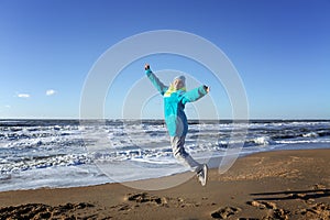 A young girl jumps on the seashore. Happy blonde woman in warm clothes in the winter season on a sunny day. Freedom and activity