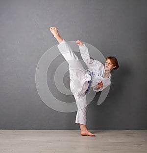 A young girl karateka in white kimono and a blue belt trains and performs a set of exercises against a gray wall