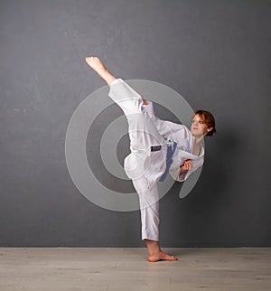 A young girl karateka in white kimono and a blue belt trains and performs a set of exercises against a gray wall