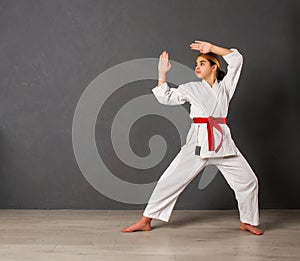 Young girl karateka in a white kimono and a red belt trains and performs a set of exercises against a gray wall