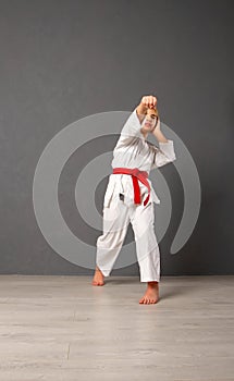 Young girl karateka in a white kimono and a red belt trains and performs a set of exercises against a gray wall