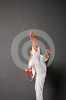 Young girl karateka in a white kimono and a red belt trains and performs a set of exercises against a gray wall