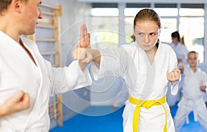 Young girl in kimono and colored belt practicing karate punch block during group martial arts lesson in gym, accompanied