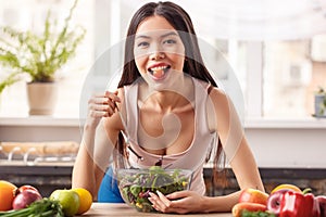 Young girl at kitchen healthy lifestyle leaning on table eating salad holding tomato in mouth looking camera playful