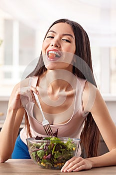 Young girl at kitchen healthy lifestyle leaning on table eating salad holding tomato with teeth looking camera cheerful