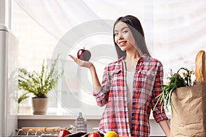 Young girl at kitchen healthy lifestyle leaning on table near bag with products looking camera curious showing apple