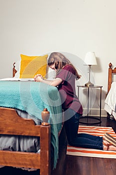 A young girl kneeling and praying to God at the side of a bed in her bedroom