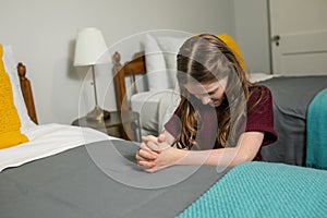 A young girl kneeling and praying to God at the side of a bed in her bedroom