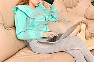 A young girl with a laptop is sitting on a leather sofa and typi