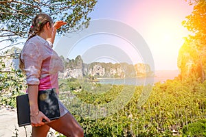 A young girl with laptop in a white shirt and pink t-shirt stands on top of a mountain overlooking a tropical beach and jungle.