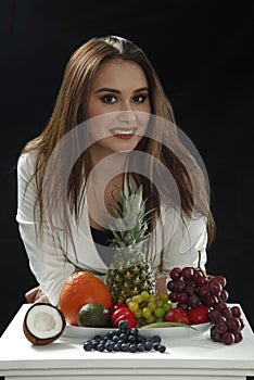 Young girl leaned on the stand and smiling close to assorti of differen fruits on black background
