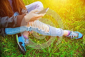 Young girl in leather jacket and ripped jeans sitting on the grass in the park and talking to friends on the phone