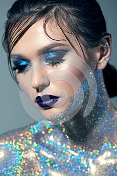 Young girl loking down with blue professional make up and wet hairstyle, with body covered a lot of blue sparkles.