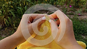 A young girl with long blond hair knits a yellow sweater in the garden in the summer. woman makes clothes with hands closeup