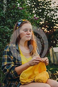 A young girl with long blond hair knits a yellow sweater in the garden in the summer. woman makes clothes with hands closeup