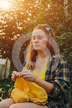 A young girl with long blond hair knits a yellow sweater in the garden in the summer. woman makes clothes with hands closeup