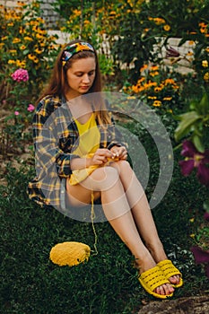 A young girl with long blond hair knits a yellow sweater in the garden in the summer. woman makes clothes with hands closeup