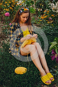 A young girl with long blond hair knits a yellow sweater in the garden in the summer. woman makes clothes with hands closeup