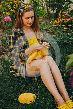 A young girl with long blond hair knits a yellow sweater in the garden in the summer. woman makes clothes with hands closeup