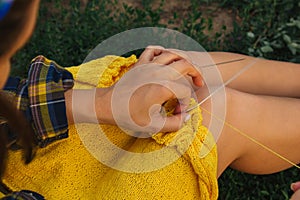 A young girl with long blond hair knits a yellow sweater in the garden in the summer. woman makes clothes with hands closeup