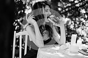 Young girl in a long evening dress is sitting at a white table in the park and eating a hamburger. Black and white photography