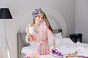 Young girl with long hair in pink bathrobe and curler on head holding iron and speaking on phone at home. She looks