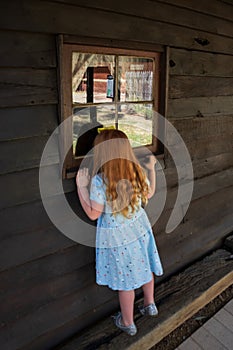A young girl with long red hair and a blue dress and upstreched arms looks curiously through a window of an old wooden building
