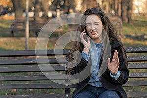 Young girl with long wavy brown hair sits on bench in park talks on phone and gesticulating. Woman in autumn park