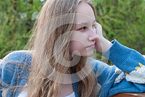 A young girl with loose blonde hair in a blue cardigan looks thoughtfully into the distance with her face propped up by