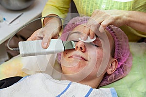 A young girl is lying on couch with a pink hat on her hair while performing an ultrasonic face cleaning with a special device for