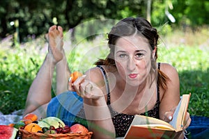 Girl with pigtails lying on the grass, reading a book and eating fruit, during a picnic in the park