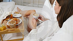 Young girl lying in light hotel apartment bed. Side view of woman in white men shirt having breakfast on small table