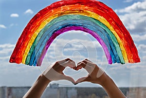 A young girl made a heart sign with her hands against the background of a painted rainbow on the window pane. bright painted rainb
