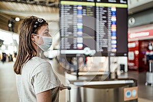 A young girl in mask waiting at the airport with  visible flight information board