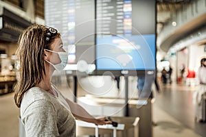 A young girl in mask waiting at the airport with  visible flight information board