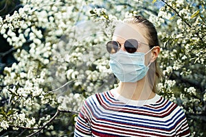 Young girl in medical mask and sunglasses on background of blossoming apple tree on summer day.