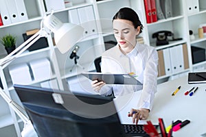 A young girl in the office holds a pen in her hand and works with documents and a computer.