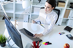 A young girl in the office holds a pen in her hand and works with documents and a computer.