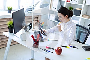 A young girl in the office holds a pen in her mouth and works with a calculator, documents and a computer.