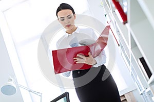 Young girl in the office near the rack and scrolls through the folder with the documents.