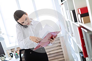 Young girl in the office near the rack and scrolls through the folder with the documents and speaks on the phone.