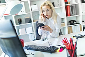 A young girl in the office sits at a desk holding a phone in her hand and talking on it through a headset.