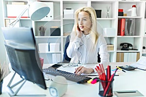 A young girl in the office sits at a table talking on the phone through a headset and working at the computer.