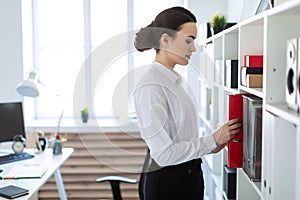 A young girl in the office is standing near the shelf and pulling out a red folder.