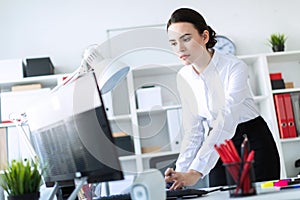 A young girl in the office is standing near the table, holding a pencil in her hand and typing text on the computer.