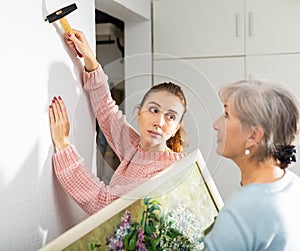 Young girl and older woman hanging painting on wall indoors